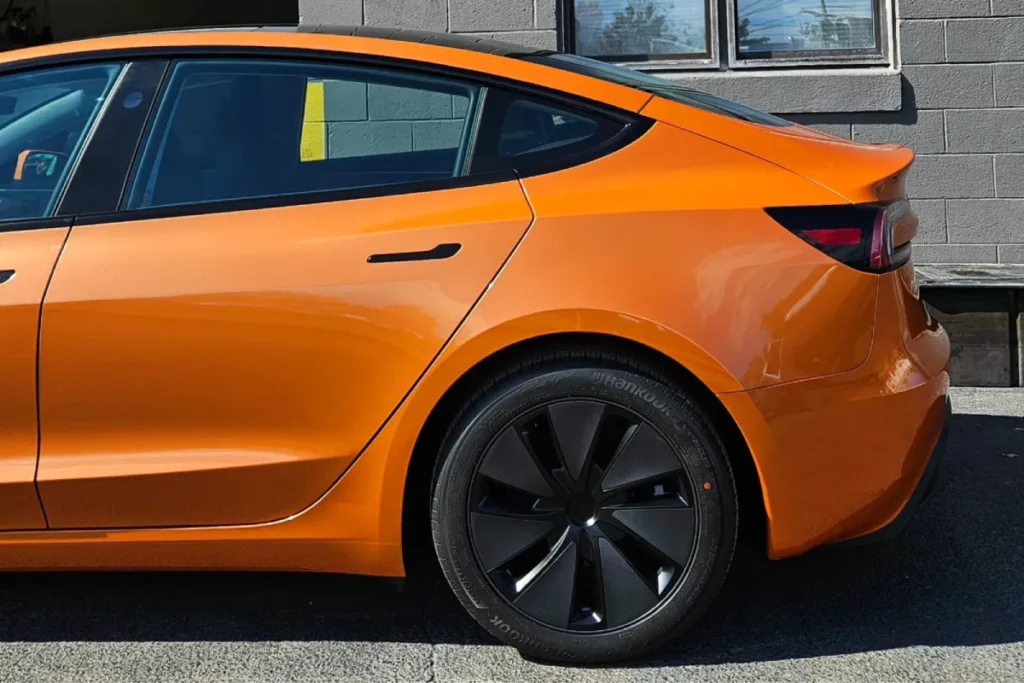 A close-up of an orange Tesla with black wheels, parked in front of a gray wall.